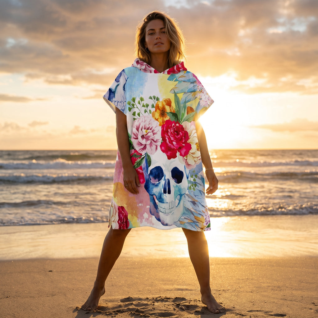 Poncho serviette à capuche motif crâne fleuri, porté par une femme posant face à l'océan au coucher du soleil.