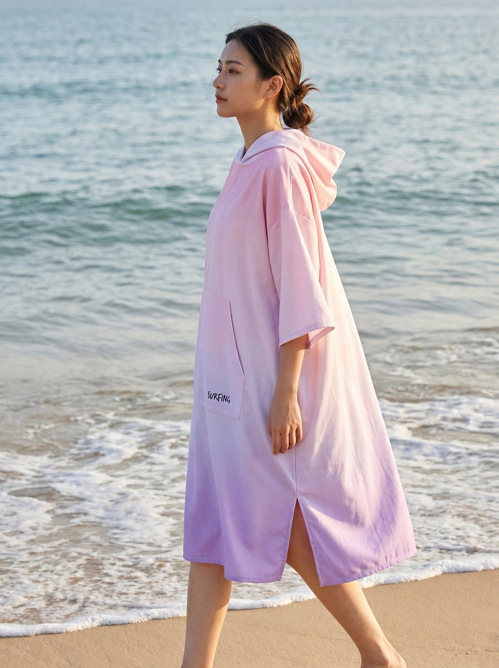 Poncho serviette à capuche motif dégradé rose, porté par une femme marchant de profil sur la plage.