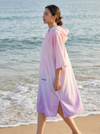 Poncho serviette à capuche motif dégradé rose, porté par une femme marchant de profil sur la plage.