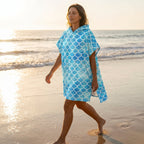 Poncho serviette à capuche motif écailles bleu ciel, porté par une femme marchant sur la plage.