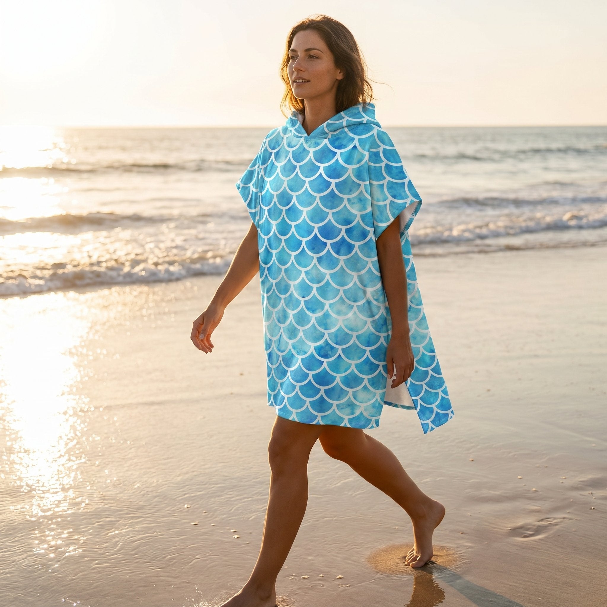 Poncho serviette à capuche motif écailles bleu ciel, porté par une femme marchant sur la plage.