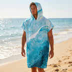 Poncho serviette a capuche motif vague marbree bleue, porte par un homme sur la plage, face a l'ocean.