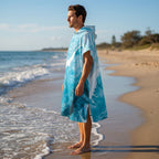 Poncho serviette a capuche motif vague marbree bleue, porte par un homme de profil, les pieds dans l'eau.