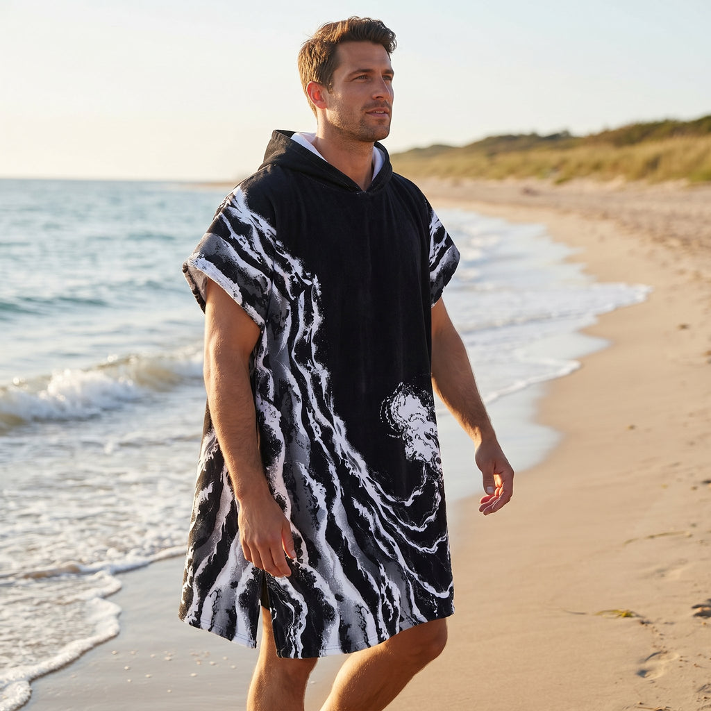 Poncho serviette a capuche motif vague noir et blanc, porté par un homme marchant de profil sur la plage.