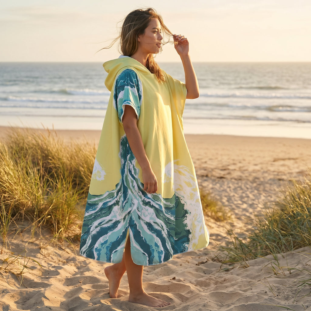 Poncho serviette à capuche motif vagues jaune pâle, porté par une femme de profil sur le sable près des dunes.