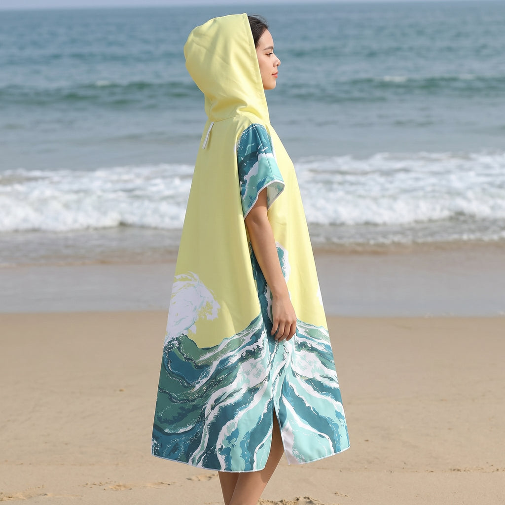 Poncho serviette à capuche motif vagues jaune pâle, porté par une femme de profil, debout sur la plage face à la mer.