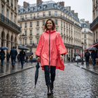 Poncho de pluie à capuche corail uni, porté par une femme souriante marchant dans une rue pavée sous la pluie.