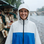 Poncho de pluie à capuche crème et bleu porté par une femme souriante sous la pluie, gros plan.