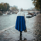 Poncho de pluie à capuche crème et bleu porté par une femme marchant de dos le long d'une rivière.