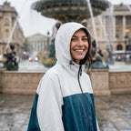 Poncho de pluie à capuche crème et bleu canard, porté par une femme souriante, gros plan devant une fontaine.