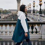 Poncho de pluie à capuche crème et bleu canard, porté de côté par une femme traversant un pont en pierre.