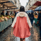 Poncho de pluie à capuche crème et corail, porté de dos par une femme marchant sous la pluie dans un marché.