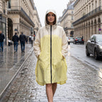 Poncho de pluie à capuche crème et jaune, porté par une femme marchant sur une rue pavée mouillée.