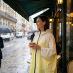 Poncho de pluie à capuche crème et jaune, porté par une femme souriante à la terrasse d'un café.