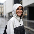 Poncho de pluie à capuche crème et noir, porté par une femme souriante, en gros plan sous la pluie.