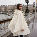 Poncho de pluie à capuche crème uni, porté par une femme souriante, de dos sur un pont, regardant par-dessus son épaule.