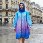 Poncho de pluie à capuche dégradé bleu et violet, porté de face par une personne marchant sur une rue mouillée.
