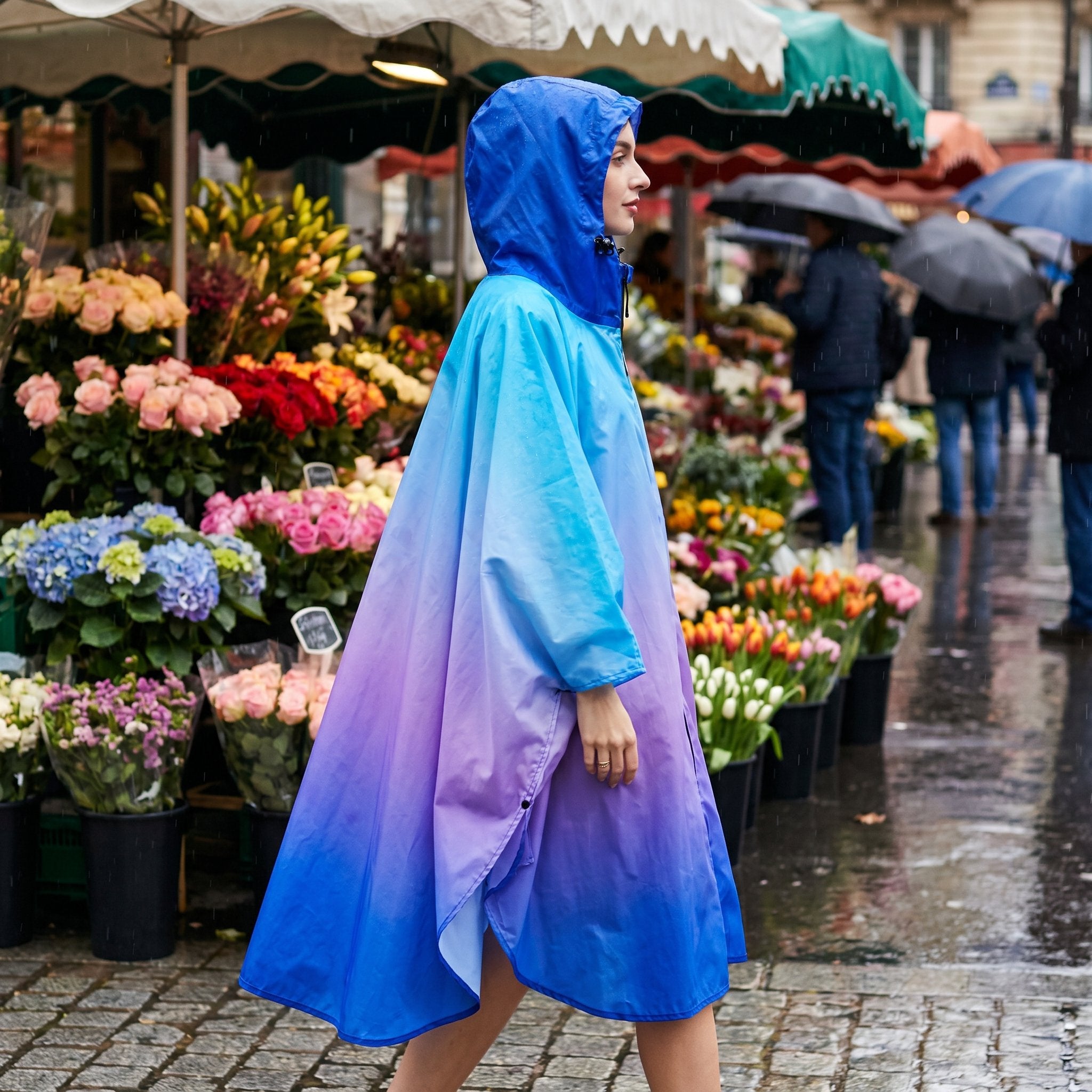 Poncho de pluie à capuche dégradé bleu et violet, porté de profil par une personne passant devant un marché aux fleurs.