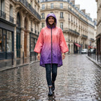 Poncho de pluie à capuche dégradé corail et violet, porté par une femme souriante marchant dans une rue pavée.
