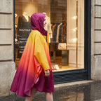 Poncho de pluie à capuche dégradé soleil, porté de côté par une femme longeant une vitrine de magasin.