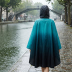 Poncho de pluie à capuche dégradé turquoise et nuit, vu de dos, marchant le long d'un canal pluvieux.