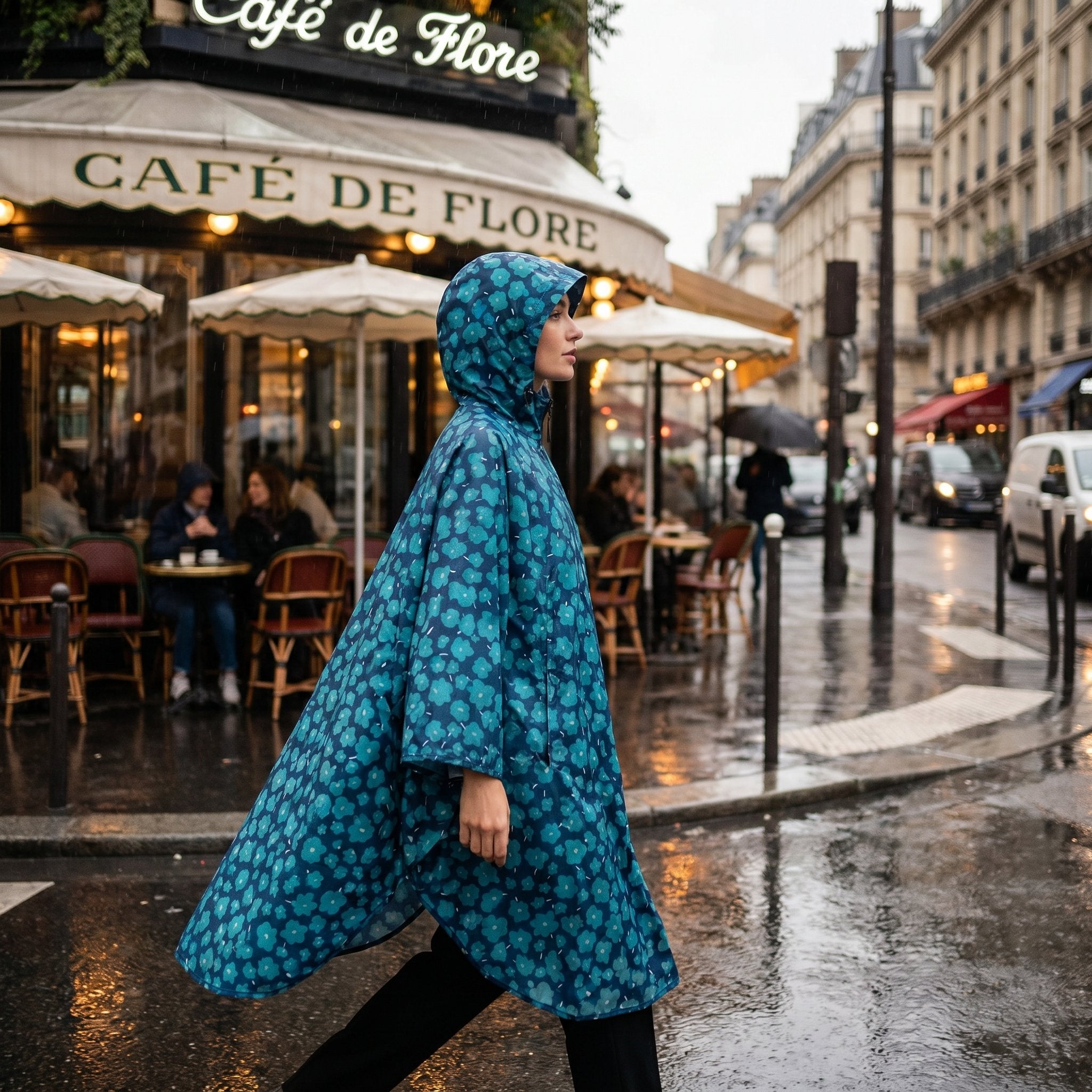 Poncho de pluie à capuche motif fleurs turquoise, une femme marche de profil sur un trottoir mouillé.