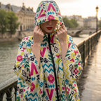 Poncho de pluie à capuche motifs multicolores, porté par une femme ajustant sa capuche, avec un pont en arrière-plan.