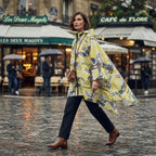 Poncho de pluie à capuche motif graphique jaune, porté par une femme traversant une rue pavée mouillée, vue de profil.