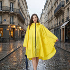 Poncho de pluie à capuche jaune uni, porté par une femme marchant sur une rue pavée mouillée.