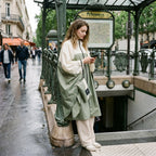 Poncho de pluie à capuche kaki et crème, porté par un modèle debout près d'une entrée de métro, regardant son téléphone.