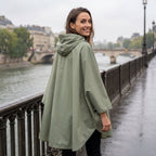 Poncho de pluie à capuche kaki uni, porté par une femme souriante, vue de dos sur un pont.