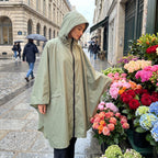 Poncho de pluie à capuche kaki uni, porté par une femme de profil devant un étal de fleurs.
