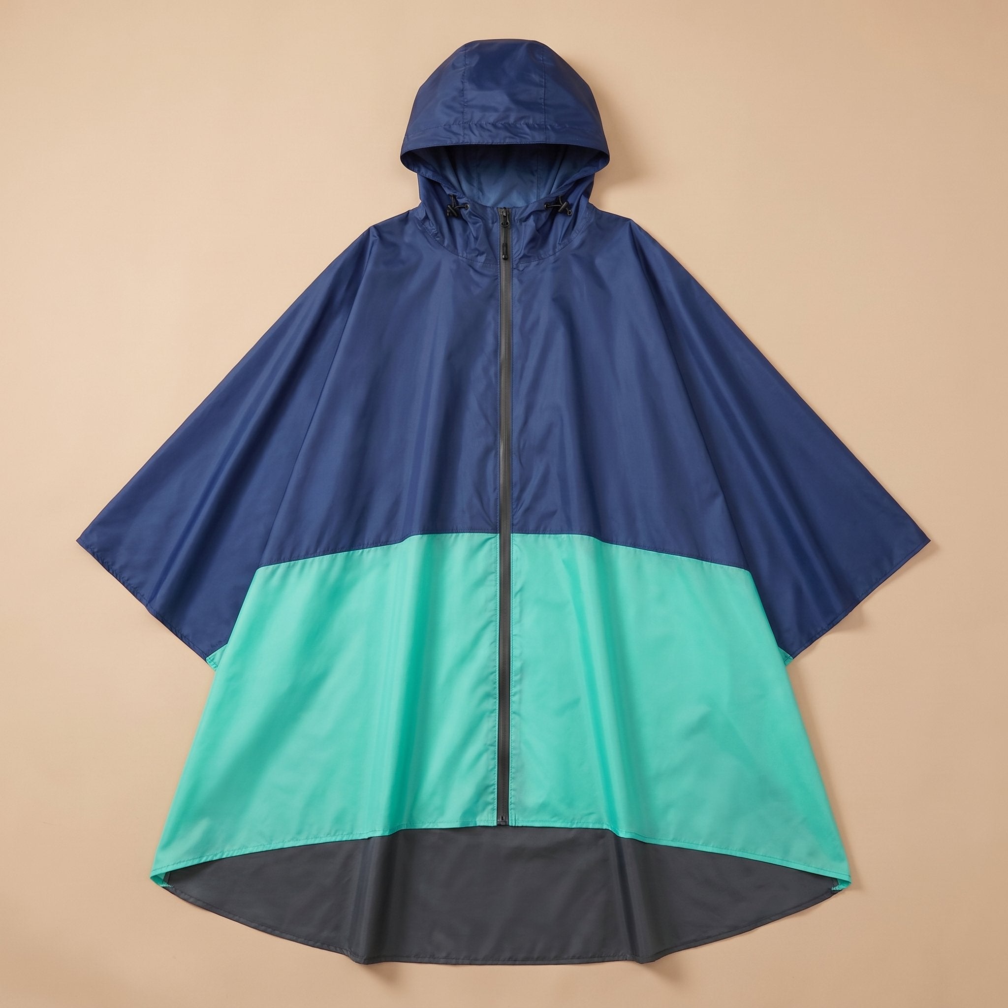 Poncho de pluie à capuche marine et menthe posé à plat sur un fond uni beige, montrant ses trois couleurs.