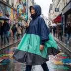 Poncho de pluie à capuche marine et menthe porté par une femme tenant un parapluie sur une rue colorée et mouillée.