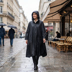 Poncho de pluie à capuche noir uni, porté par une femme souriante marchant sur un trottoir.