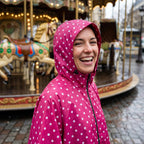 Poncho de pluie à capuche pois fuchsia, porté par une femme souriante, avec un manège en arrière-plan.