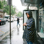 Poncho de pluie à capuche motif Pois jaunes, porté par une femme vérifiant sa montre à un arrêt de bus.