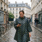 Poncho de pluie à capuche motif Pois jaunes, porté par une femme marchant dans une rue pavée et regardant son téléphone.