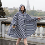Poncho de pluie à capuche rayures marines, porté par une femme souriante marchant sur un pont, tenant une tasse.