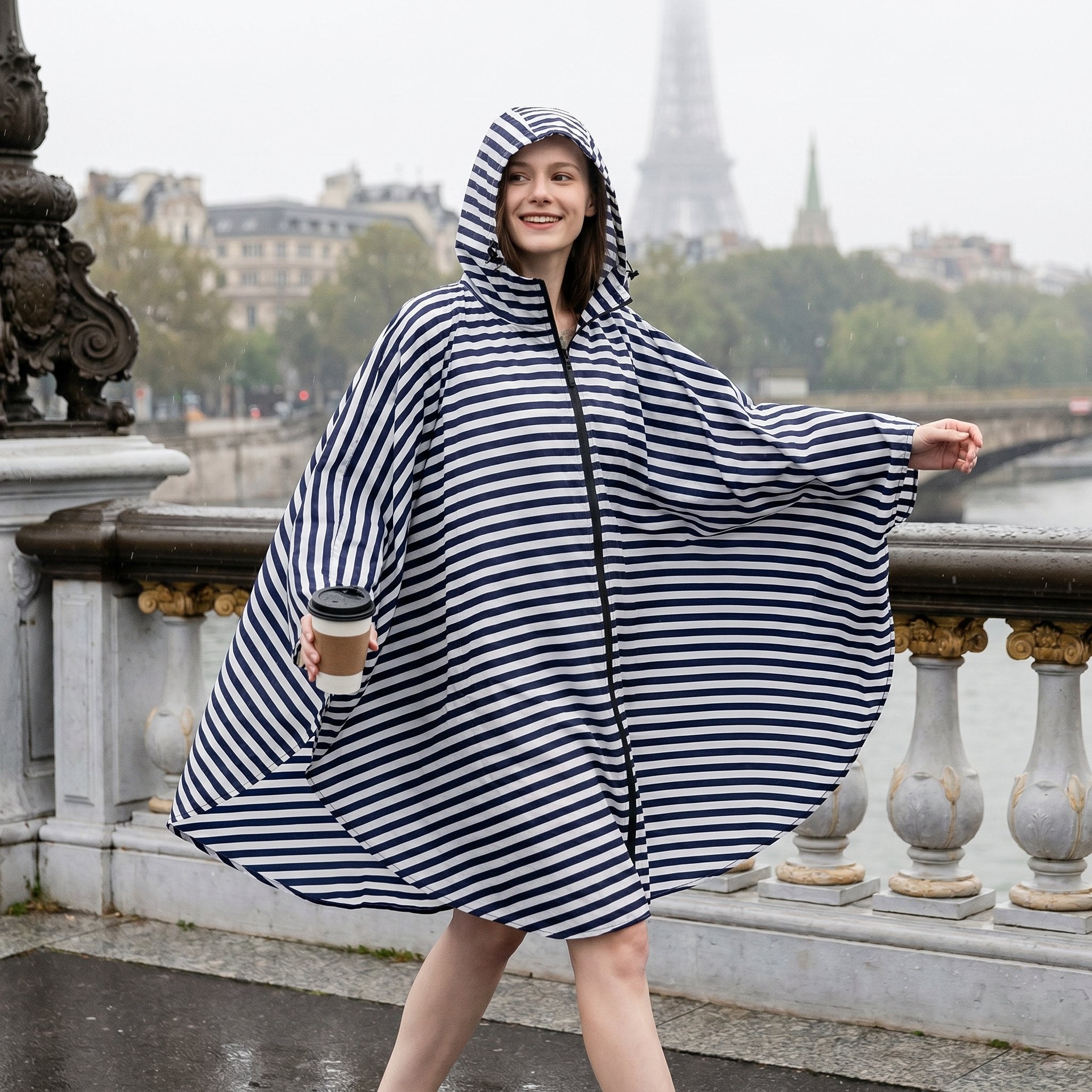 Poncho de pluie à capuche rayures marines, porté par une femme souriante marchant sur un pont, tenant une tasse.