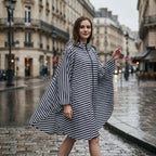 Poncho de pluie à capuche rayures marines, porté par une femme marchant sur une rue pavée mouillée en ville.