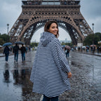 Poncho de pluie à capuche rayures marines, porté par une femme de dos regardant par-dessus son épaule, devant un monument.