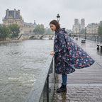 Poncho de pluie à capuche motif roses marine, porté par une femme sur un pont, regardant le fleuve.