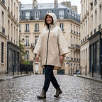 Poncho de pluie à capuche sable uni, porté par une femme souriante marchant sur une rue pavée mouillée.