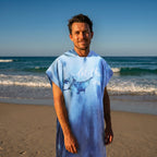 Poncho serviette à capuche motif raie manta, porté par un homme souriant, cadrage mi-corps sur la plage.