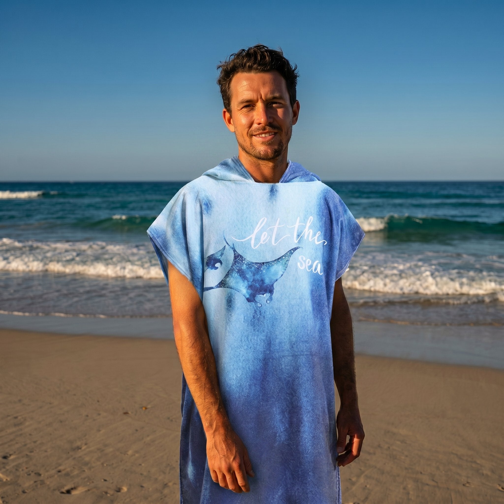 Poncho serviette à capuche motif raie manta, porté par un homme souriant, cadrage mi-corps sur la plage.