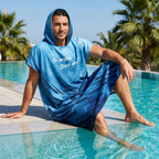 Poncho serviette à capuche motif baleine et lune, porté par un homme assis au bord d'une piscine, les pieds dans l'eau.