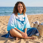 Poncho serviette à capuche motif dauphin turquoise, porté par une femme souriante assise en tailleur sur le sable.