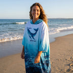 Poncho serviette à capuche motif dauphin turquoise, porté par une femme souriante, debout face à l'océan.