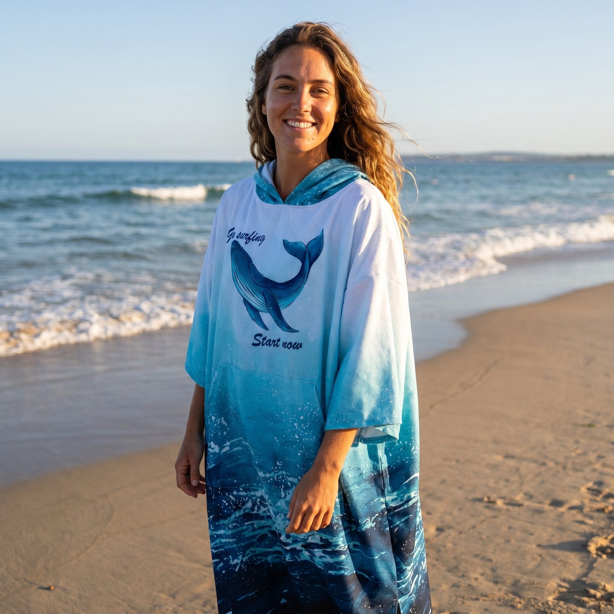 Poncho serviette à capuche motif dauphin turquoise, porté par une femme souriante, debout face à l'océan.